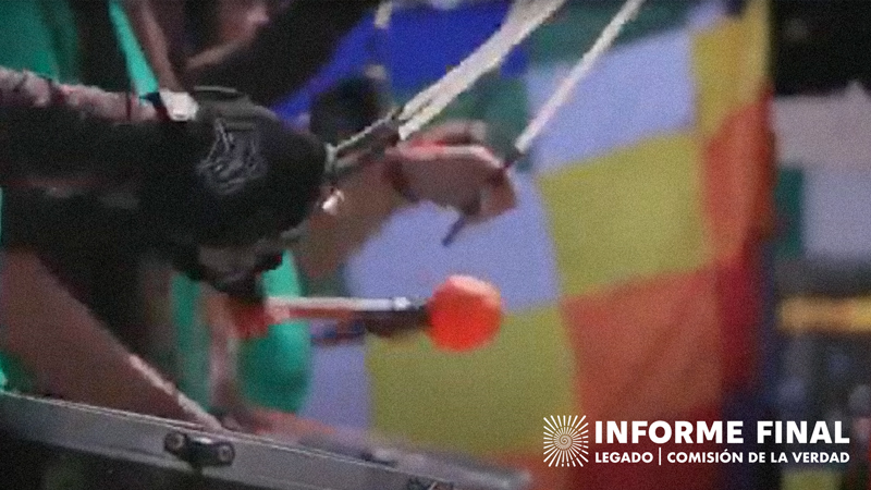 Detalle de manos tocando surdos (instrumentos de percusión) al fondo una bandera Wiphala