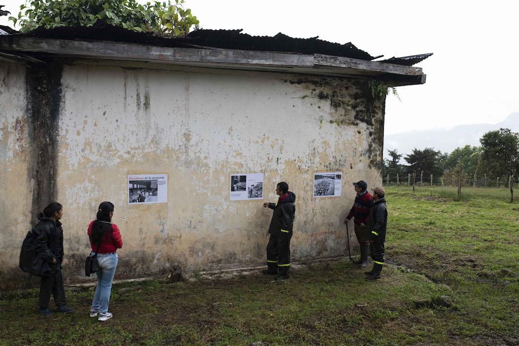 5 Personas observan afiches pegados a los muros en la fachada de una edificación abandonada. vegetación y cielo nublado