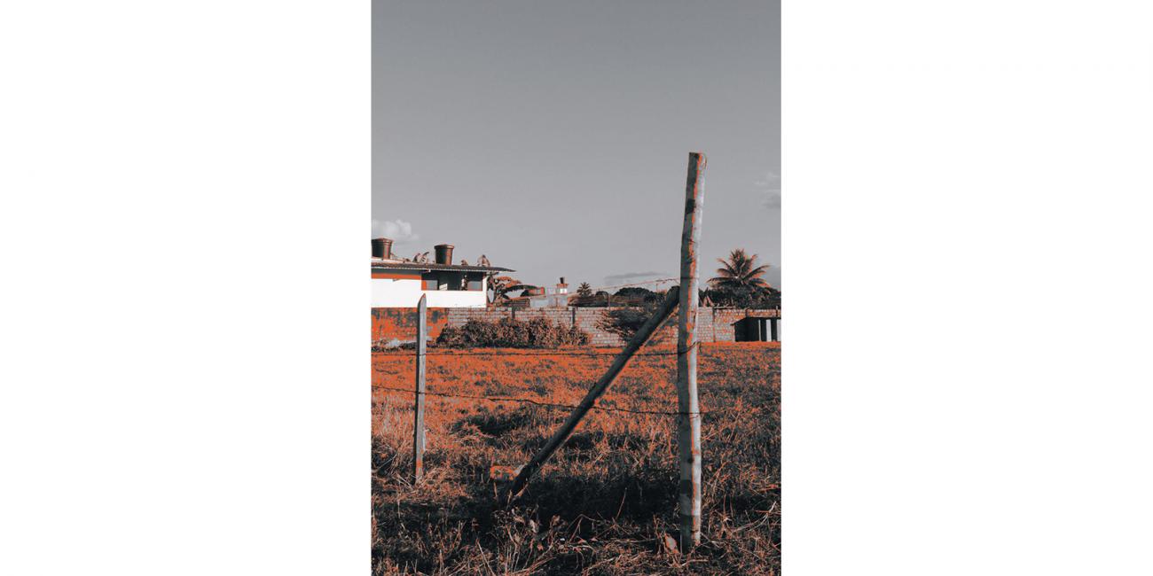 Cerca de alambre de púas. Terreno baldío, al fondo, muro de ladrillos y casa